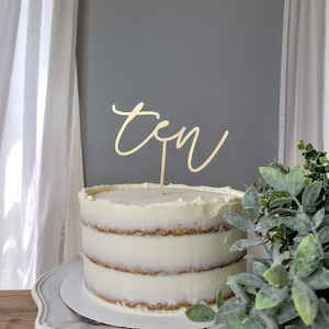 May include: A white cake with vanilla frosting and a gold 'ten' cake topper. The cake is on a white cake stand and there is a green plant in the background.