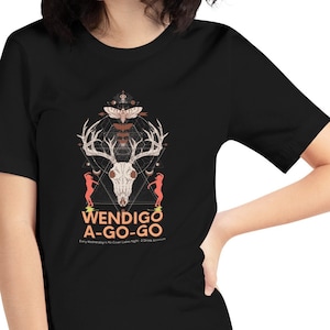 Op de afbeelding: Zwart T-shirt met een grafisch ontwerp met een herten schedel, geweien en de tekst "WENDIGO A-GO-GO". Het ontwerp bevat geometrische vormen en figuren in tinten wit, oranje en rood. Het shirt heeft een ronde hals en korte mouwen.