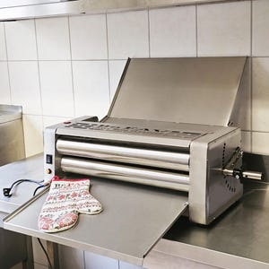 May include: A stainless steel pasta roller machine with a lever and a removable tray. The machine has a red power switch and a white oven mitt is on the tray.