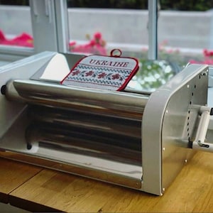 May include: A white and silver pasta maker with a red and white patterned potholder on top. The potholder has the word "UKRAINE" embroidered on it.
