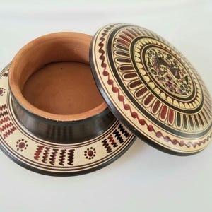 May include: A decorative, open ceramic box with a lid. The lid and base feature intricate patterns in black, red, and beige. The lid's design includes a central image and a border of repeating shapes. The interior is a natural terracotta color.
