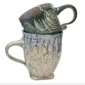 May include: Two stacked ceramic mugs. The top mug is green with a dark rim and embossed spiral patterns. The bottom mug is light gray with a blue-tinted rim and features intricate, etched floral and geometric line designs across its surface.