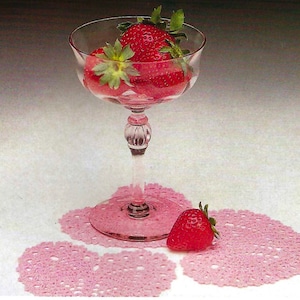 Peut inclure: Un gobelet en verre transparent rempli de fraises fra&icirc;ches, pos&eacute; sur un napperon rose en crochet. Plusieurs autres napperons roses en crochet sont dispers&eacute;s autour du gobelet, et une seule fraise est &agrave; proximit&eacute;. Le gobelet a une tige d&eacute;corative.