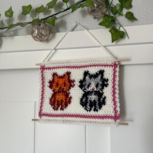 May include: A white crocheted wall hanging with a pink border featuring two cats. One cat is orange and the other is gray and white.