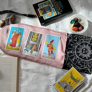May include: A pink and black tarot bag with a black and white zodiac design. The bag is open and shows a spread of tarot cards, including the Three of Cups, the Three of Swords, and the Two of Wands. A book titled "A Complete Guide to the Tarot" by Eden Gray is visible in the background.
