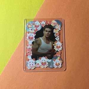 Puede incluir: Un portafotos de acrílico transparente con una foto de un hombre con una camiseta sin mangas blanca. La foto está rodeada de pegatinas decorativas de personajes de dibujos animados con lazos y pasteles. El fondo es una mezcla de amarillo y naranja.