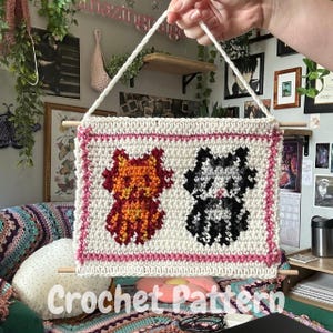 May include: A white crochet wall hanging with a pink border featuring two pixelated cats. One cat is orange and the other is gray. The hanging is attached to a wooden dowel rod with white yarn.