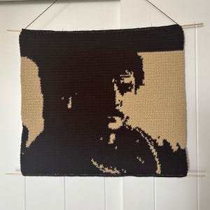 Puede incluir: Un tapiz de pared de ganchillo que presenta un retrato de una persona con cabello oscuro y una camisa oscura. El fondo es de color beige claro.