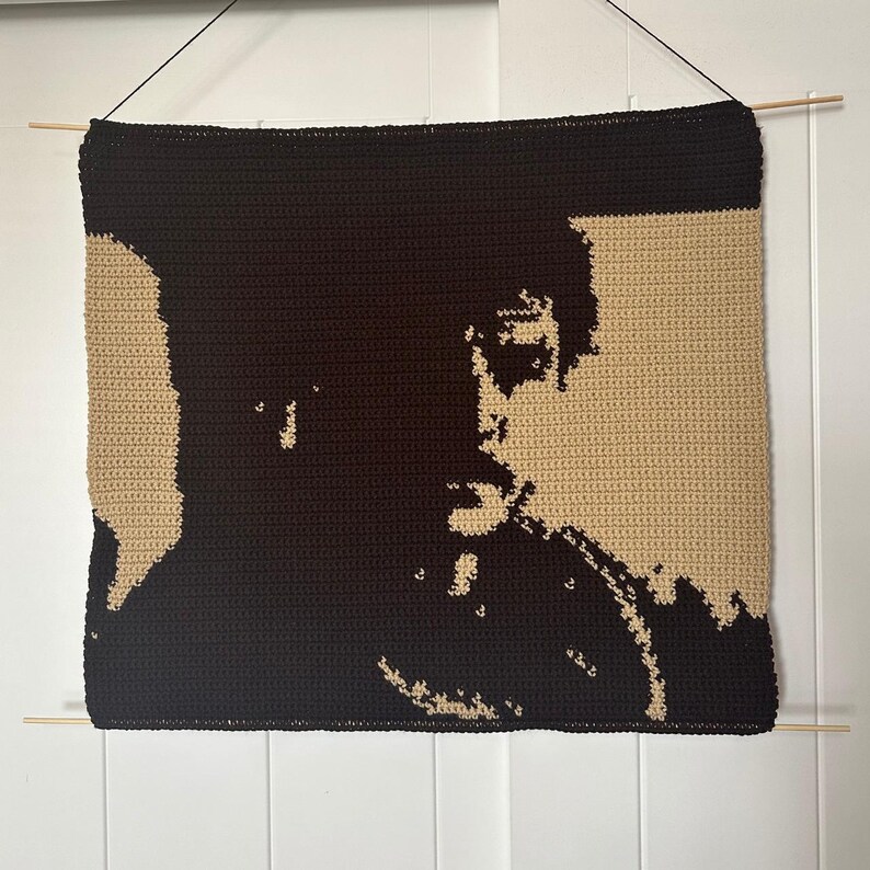 Puede incluir: Un tapiz de pared de ganchillo que presenta un retrato de una persona con cabello oscuro y una camisa oscura. El fondo es de color beige claro.