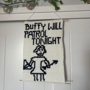 May include: White crocheted wall hanging with black text that reads "Buffy Will Patrol Tonight" and a black and white pixelated image of a person holding a stake.