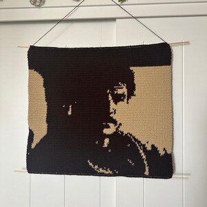Puede incluir: Un tapiz de pared tejido a crochet con la silueta de una cara en hilo negro sobre un fondo beige.