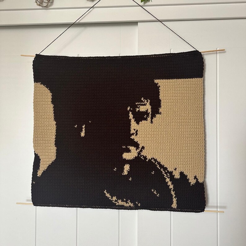 Puede incluir: Un tapiz de pared tejido a crochet con la silueta de una cara en hilo negro sobre un fondo beige.