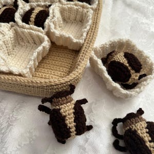 May include: A crocheted basket with beige and white yarn, containing crocheted brown and beige bugs. The bugs are sitting in the basket and on a white surface.