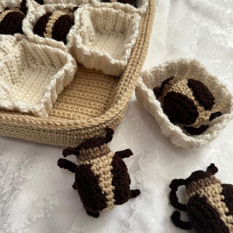 May include: A crocheted basket with beige and white yarn, containing crocheted brown and beige bugs. The bugs are sitting in the basket and on a white surface.