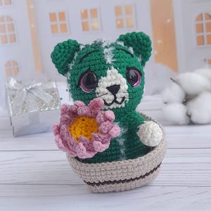 May include: A green crocheted cactus toy holding a pink flower. The cactus is wearing a white crocheted face with pink eyes and a white nose. The cactus is sitting in a brown crocheted pot.