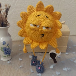 Puede incluir: Un sol amarillo de crochet con una cara somnolienta, usando pantuflas de conejo grises y sosteniendo una taza de café.