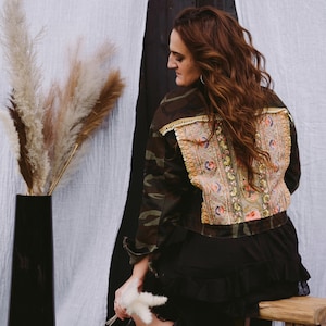 Könnte beinhalten: Tarnjacke mit einem bestickten Rückenteil mit Blumen- und Paisley-Designs in Rosa, Gold und Blau. Die Jacke hat goldfarbene Verzierungen und wird mit einem schwarzen Rock kombiniert. Ein einzigartiges Modeaccessoire.