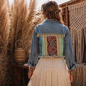 Puede incluir: Una chaqueta de mezclilla azul con un panel trasero de bordado rojo, verde y dorado, acentuado por una fila de cuentas blancas colgantes en el borde superior. La chaqueta tiene mangas largas, cuello y un dobladillo deshilachado en la parte inferior.