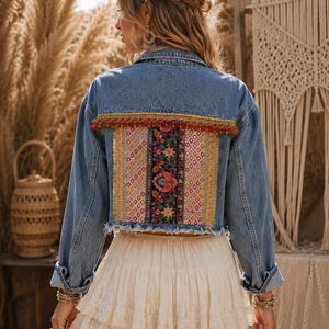 May include: A blue denim jacket featuring a back panel with a central black floral embroidery strip, flanked by gold metallic patterned fabric and a red tassel fringe trim. The jacket has a collar, long sleeves, and a frayed hemline at the waist.