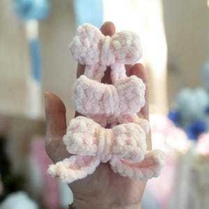 May include: Three pink fluffy yarn bows. The bows are tied together and are held in a hand.