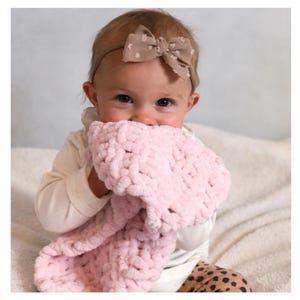 May include: A baby wearing a beige bow headband and a white long-sleeved top, holding a soft, light pink, chunky knit blanket. The baby is looking directly at the camera. The baby is wearing patterned trousers.