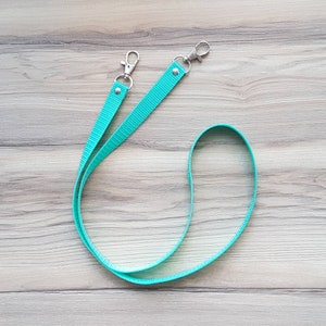 Könnte beinhalten: Ein türkisfarbenes Lanyard mit zwei silbernen Drehclips. Das Lanyard ist aus gewebtem Material und etwa 76 cm lang.