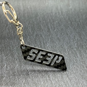 May include: A silver keychain with a black and white carbon fiber design. The keychain has a silver ring and a silver clip. The carbon fiber design has the text "SE3P" in white.