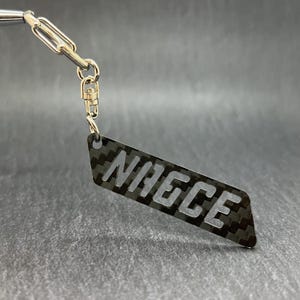 以下が含まれることがあります： 白い文字で「NICE」と書かれた黒いカーボンファイバー製のタグが付いたシルバーのキーホルダーです。