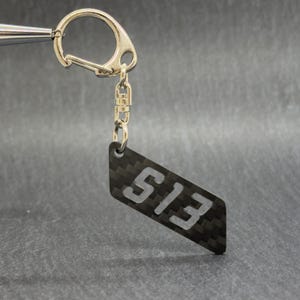 Könnte beinhalten: Ein schwarzer Schlüsselanhänger aus Kohlefaser mit der weißen Nummer "S13". Der Schlüsselanhänger hat einen silbernen Verschluss und eine kleine Kette. Der Hintergrund ist dunkelgrau.