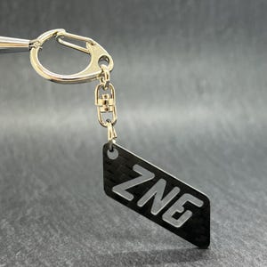 以下が含まれることがあります： 白い文字で「ZNE」と書かれた、黒と銀の四角いタグが付いた銀色のキーチェーンです。