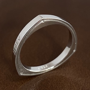May include: A silver ring with a square shape and a small diamond set in the top. The ring is engraved with the word "JEREV".
