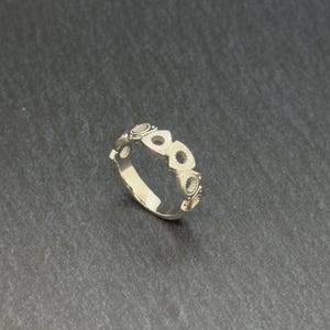 May include: A silver ring featuring a repeating pattern of stylized, openwork shapes. The ring has a polished finish, reflecting light. The design includes a series of geometric elements, creating a modern and unique aesthetic. The ring is shown on a dark gray surface.