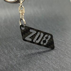 Op de afbeelding: Een zwarte carbon fiber sleutelhanger met het woord "ZIJI3" uitgesneden in wit.