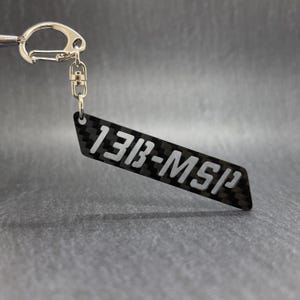 May include: A black and gray carbon fiber keychain with the text "13B-MSP" in white. The keychain has a silver metal clasp and key ring.