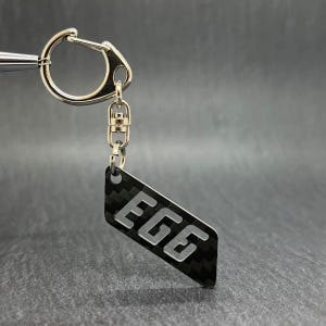 May include: A silver keychain with a black and gray carbon fiber-style tag. The tag is shaped like a parallelogram and features the word "EGG" in a bold, sans-serif font. The keychain has a silver clasp.