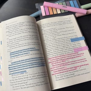 May include: An open paperback book with printed text, featuring multiple lines highlighted in blue and pink ink. The pages are marked with small, colourful adhesive index tabs along the outer edge, and several highlighters are placed nearby.