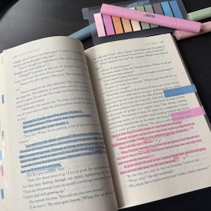 Könnte beinhalten: Ein aufgeschlagenes Taschenbuch mit gedrucktem Text, bei dem mehrere Zeilen mit blauer und rosa Tinte markiert sind. Die Seiten sind am äußeren Rand mit kleinen, bunten Klebe-Indexreitern versehen, und mehrere Textmarker liegen daneben.