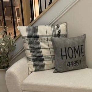 May include: A cream-colored bench with two decorative pillows. One pillow has a black and white plaid pattern, and the other is gray with the word "HOME" and the number "44857" printed on it. The bench is in front of a staircase.