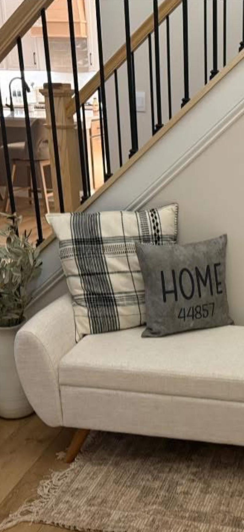 May include: A cream-colored bench with two decorative pillows. One pillow has a black and white plaid pattern, and the other is gray with the word "HOME" and the number "44857" printed on it. The bench is in front of a staircase.