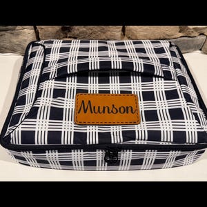 May include: A black and white plaid travel bag with a brown leather patch that reads "Munson". The bag has a black zipper and a handle on top. The bag is rectangular and appears to be made of a durable material.