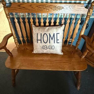 May include: A wooden bench with a high back and armrests. The back of the bench has a decorative carved design. A rectangular pillow with the word "HOME" and the number "43410" is on the seat.