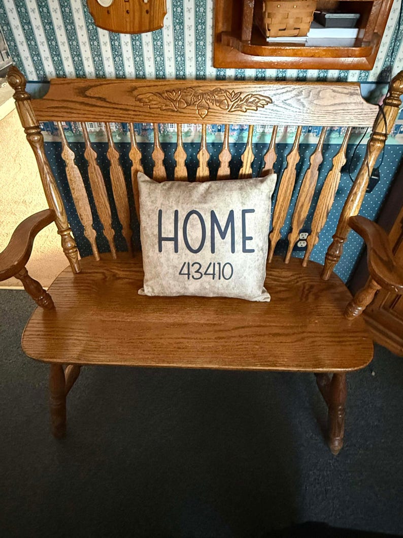 May include: A wooden bench with a high back and armrests. The back of the bench has a decorative carved design. A rectangular pillow with the word "HOME" and the number "43410" is on the seat.