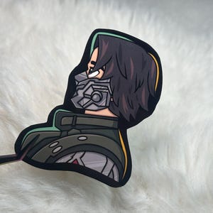 May include: A die-cut sticker featuring a profile illustration of a character with dark hair, wearing a grey mechanical face mask and a dark green high-collared jacket with silver accents. The design has a thick black border and a thin green outline.