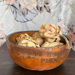 May include: A rustic, round wooden bowl with a distressed orange and brown finish, filled with dried, beige-colored flowers. The bowl sits on a dark surface, with a floral patterned fabric in the background.