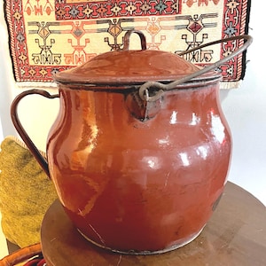 May include: A large, red, enameled, metal pot with a lid and a wire handle. The pot has a rounded body and a wide opening. The pot is sitting on a wooden table.