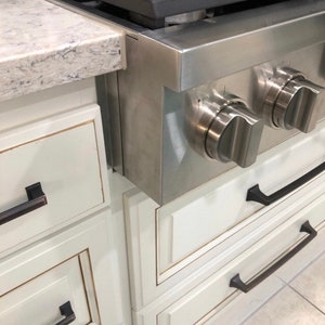 May include: A stainless steel gas hob with two large control knobs. The hob is installed above white kitchen cabinets with black handles. A granite worktop is visible on the left.