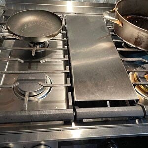May include: Stainless steel hob with a frying pan, a large pot, and a rectangular metal cover. The hob has four burners and is made of brushed metal. The frying pan is black and round, and the pot is copper-coloured.