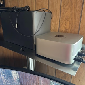 May include: A silver Apple Mac Mini computer with the Apple logo on top, and a black speaker are mounted on a metal shelf. Cables are connected to the devices. The shelf is mounted on a wooden wall.