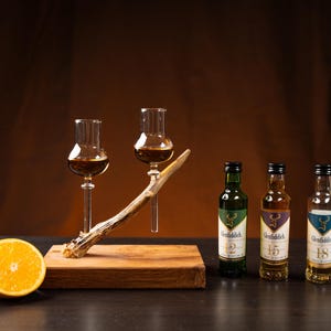 Puede incluir: Tres botellas pequeñas de whisky escocés de malta Glenfiddich, etiquetadas con 12, 15 y 18 años de edad, se muestran en un soporte de madera con una decoración de asta de ciervo. Dos decantadores de vidrio transparente con líquido marrón también están en el soporte. Dos naranjas están en primer plano.