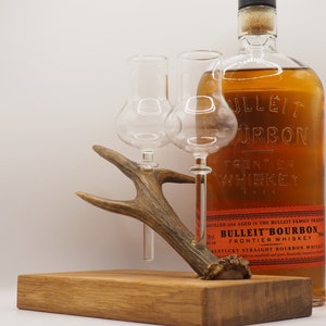 Op de afbeelding: Een houten basis met een set hertengewei-decanters, die twee heldere glazen decanters bevatten. Op de achtergrond staat een fles Bulleit Bourbon Frontier Whiskey.